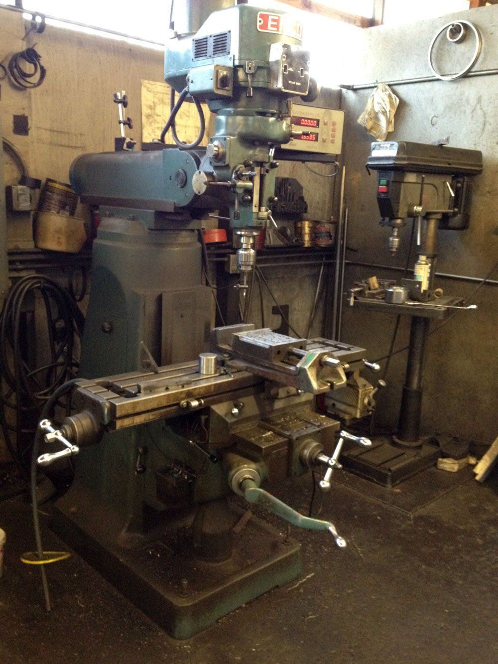 Vertical milling machine in a workshop, dark green with a rectangular metal workpiece clamped in place.