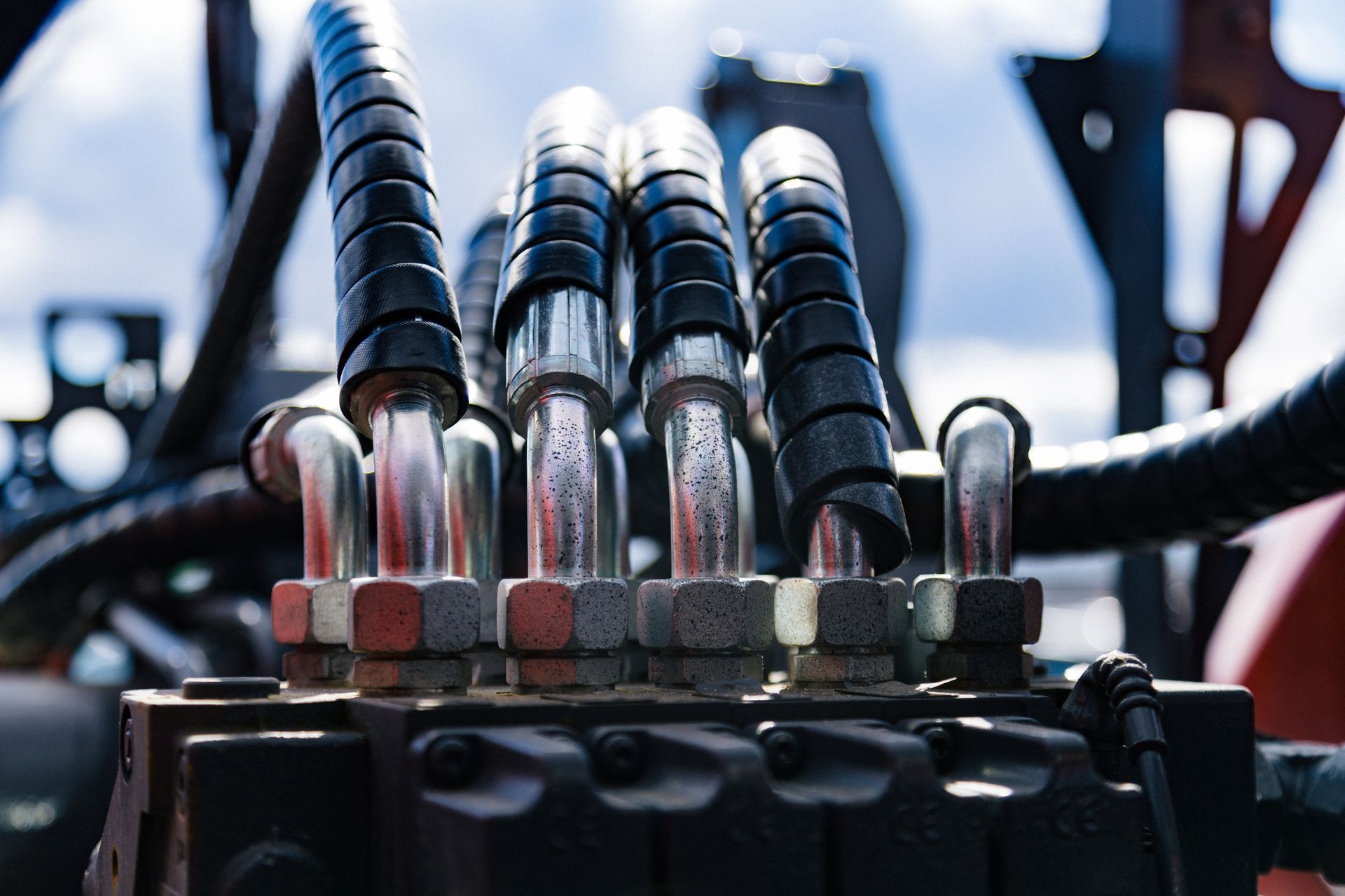 Close-up of hydraulic hoses and fittings on a dark machine, against a blurred sky background.