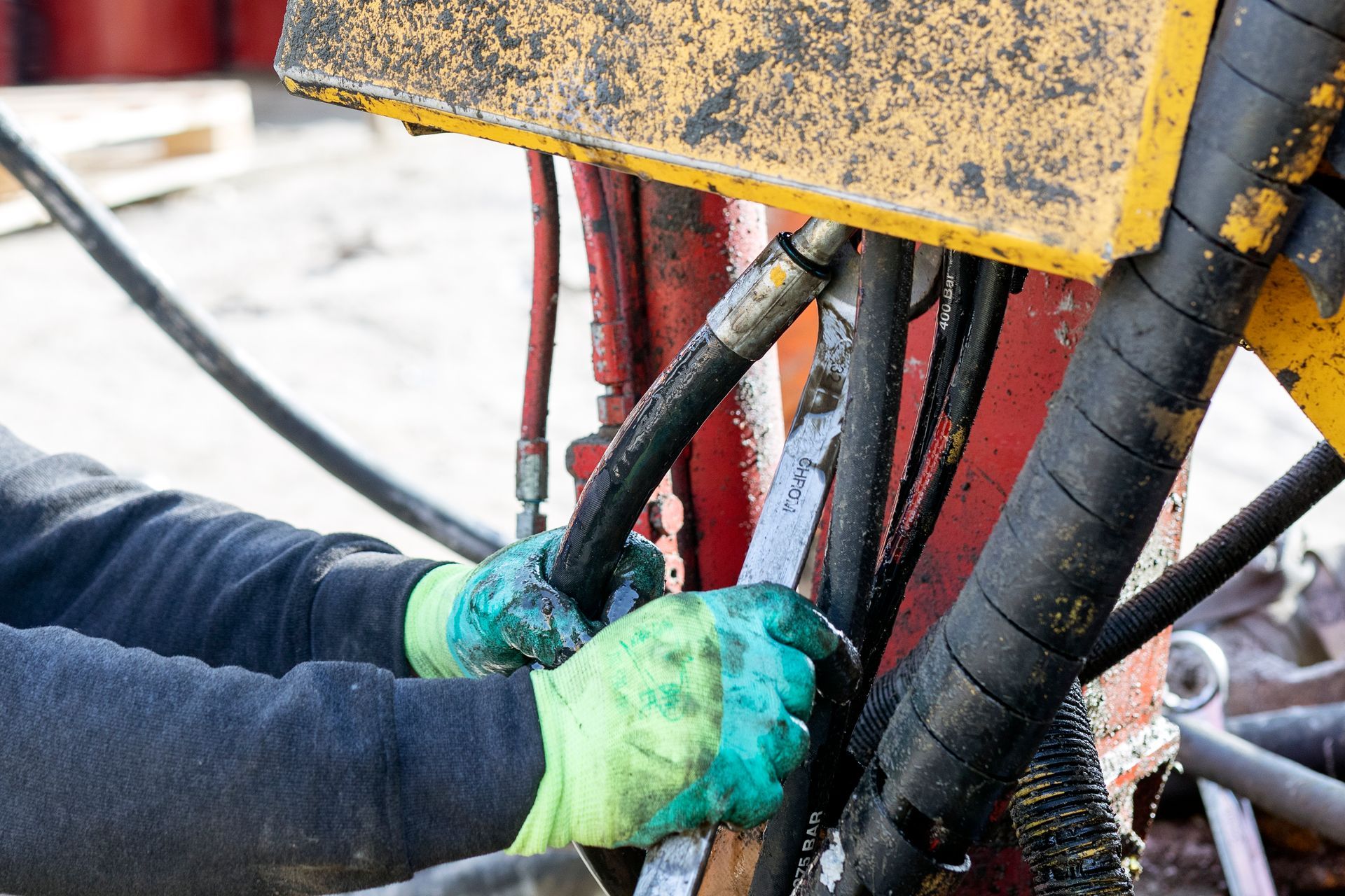 Hands wearing green gloves, working on hydraulic hoses of a machine, likely repairing or connecting them.