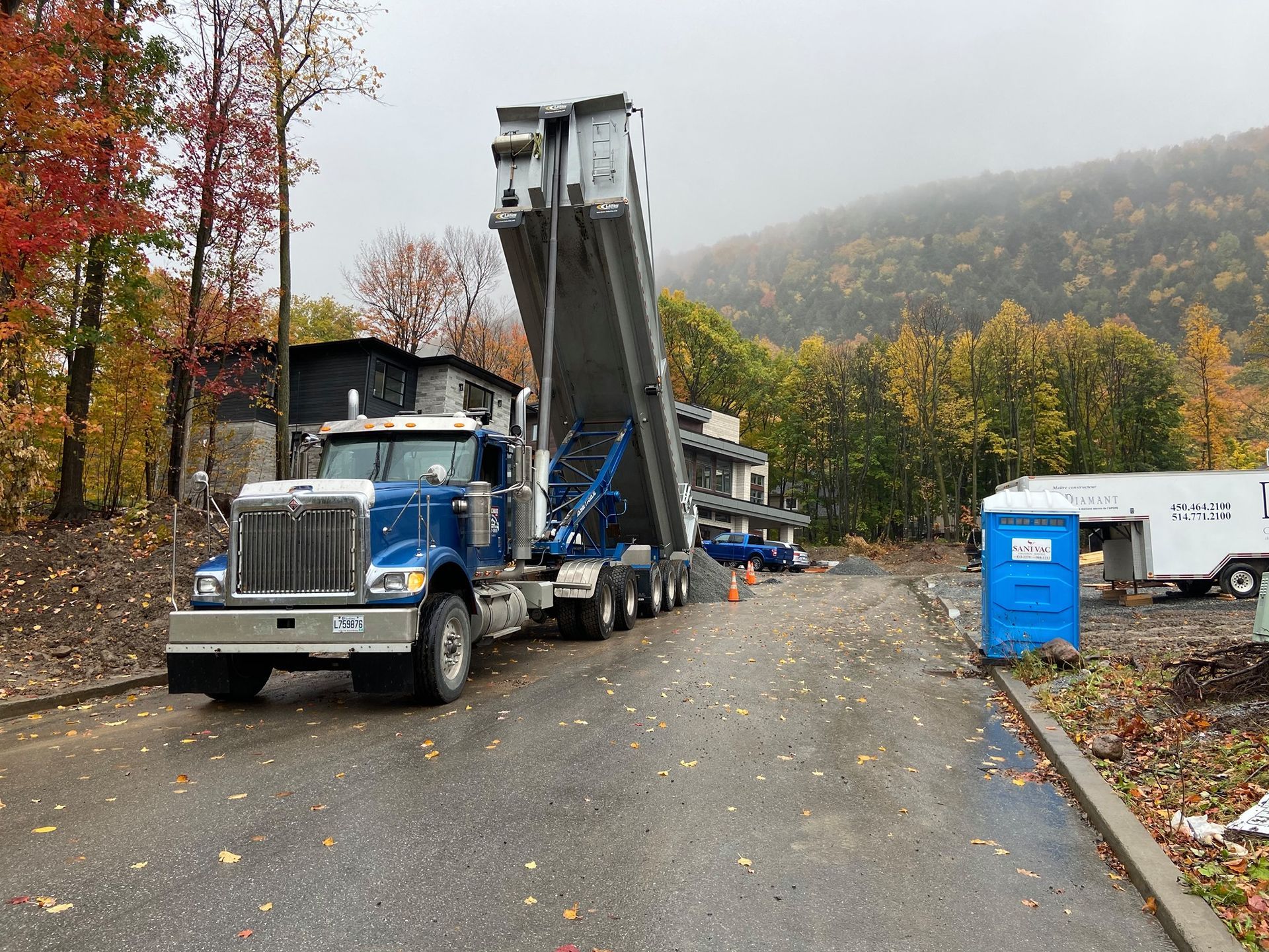Un semi-remorque bleu tractant une remorque surélevée circule sur une route de montagne détrempée en automne, avec des toilettes portables à proximité.