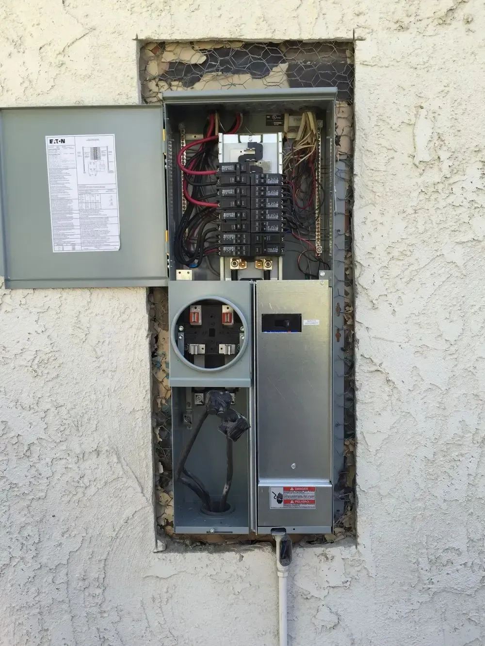 Open electrical panel recessed in a stucco wall with a visible meter box and circuit breakers.