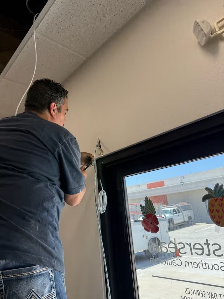 Person on ladder installing electrical wiring near a window with storefront visible outside.