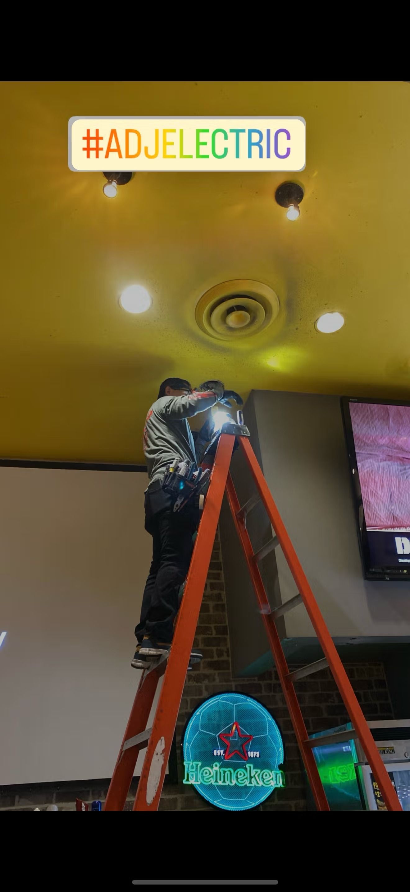 A person on a ladder working on ceiling lights, with a neon Heineken sign below.
