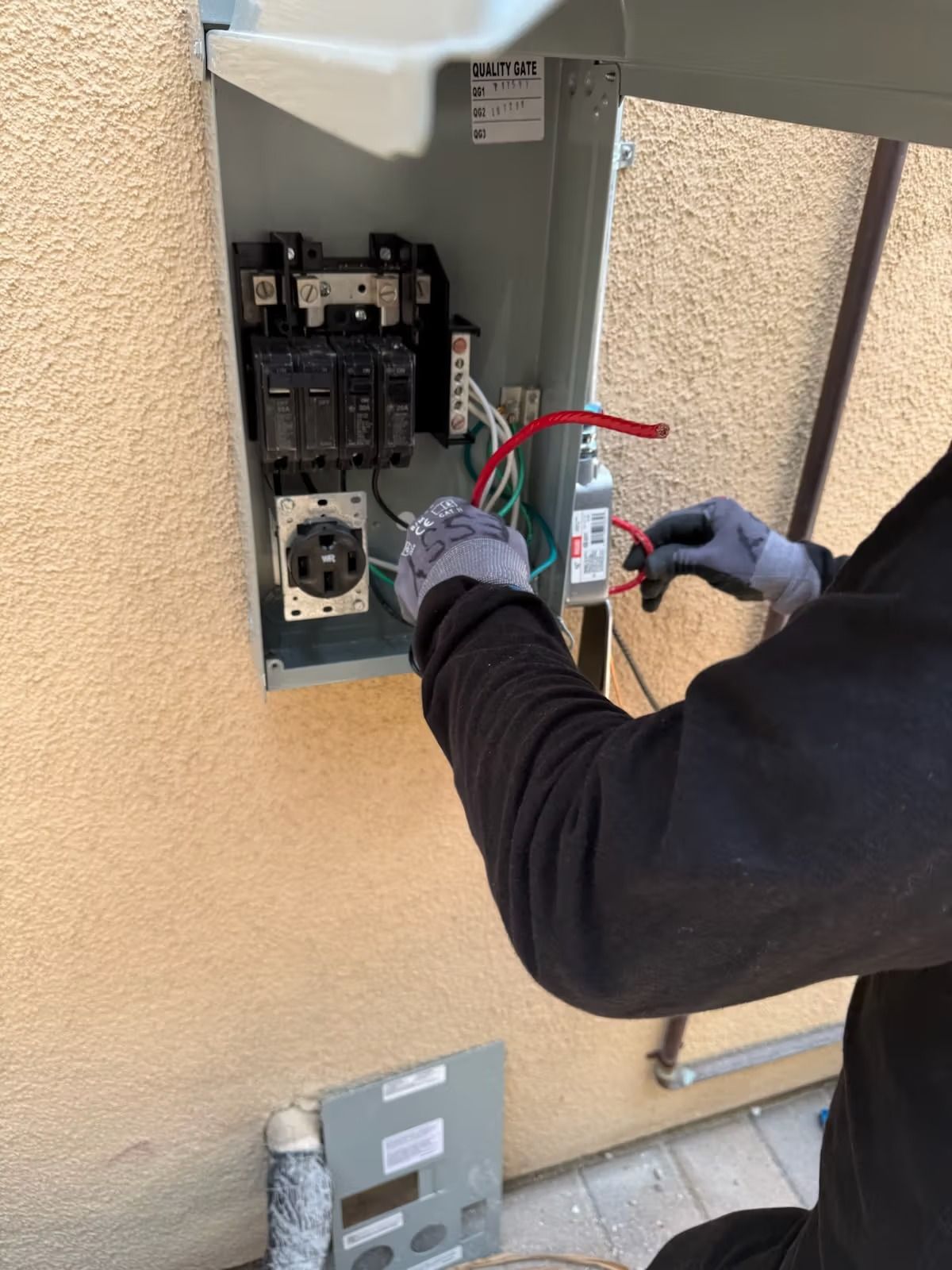 Person in gloves wiring electrical panel outdoors. Red and green wires are visible.