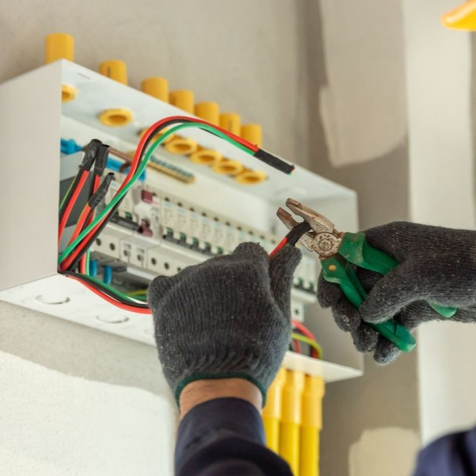Electrician using pliers on wires inside an electrical panel box, wearing gloves.