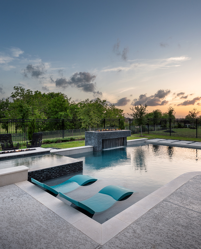 Modern backyard pool with built-in loungers at sunset, surrounded by greenery and a black fence