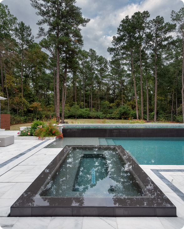 Outdoor pool with a raised black stone spa in the foreground and tall pine trees beyond.