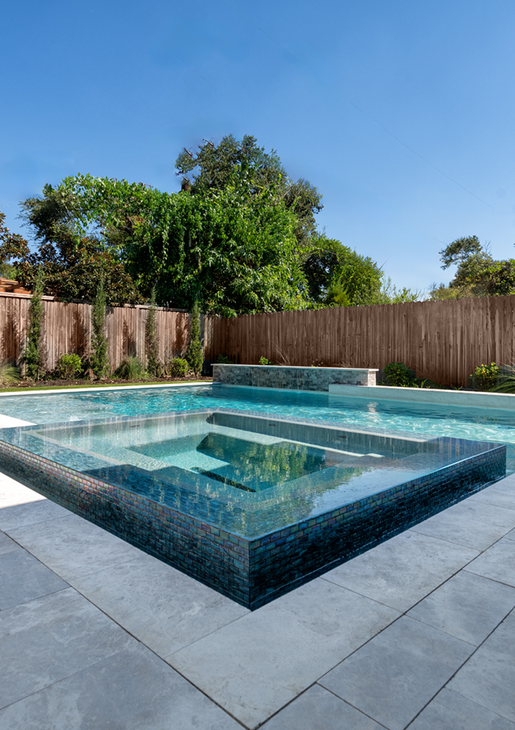 Modern backyard pool with raised square spa, light stone deck, and wood fence under a blue sky