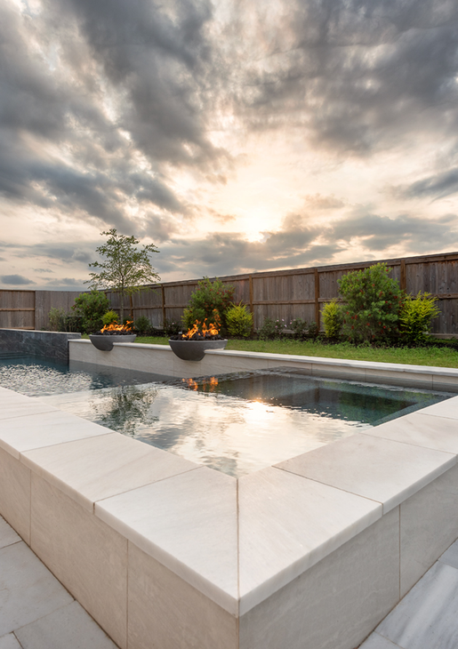 Modern backyard pool reflecting a cloudy sunset, with white stone edges and potted plants along a wooden fence