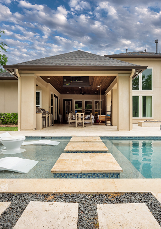 Modern backyard pool with stone stepping pads leading to a covered outdoor patio under a cloudy sky