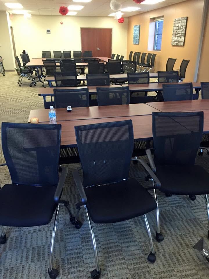 Empty conference room with rows of black chairs and tables, carpeted floor, and windows along the wall.