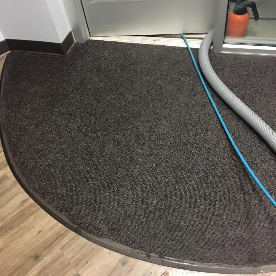 Curved dark carpeted floor mat beside wood flooring, with a blue cable and vacuum hose nearby.