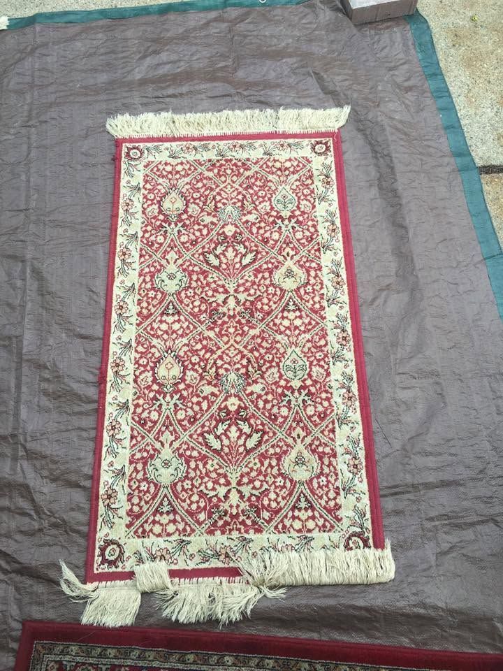 Red patterned prayer rug with tassels laid on a gray cloth surface.