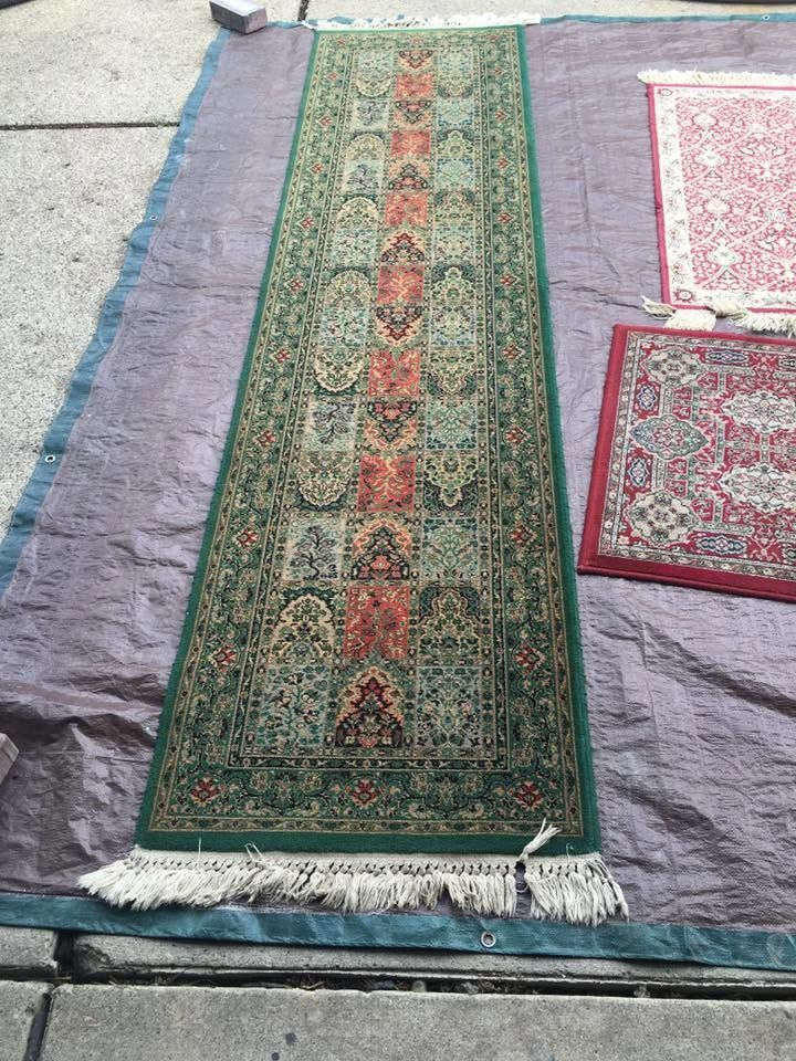 Long green patterned runner rug with red center on a gray floor, next to smaller pink-red rugs