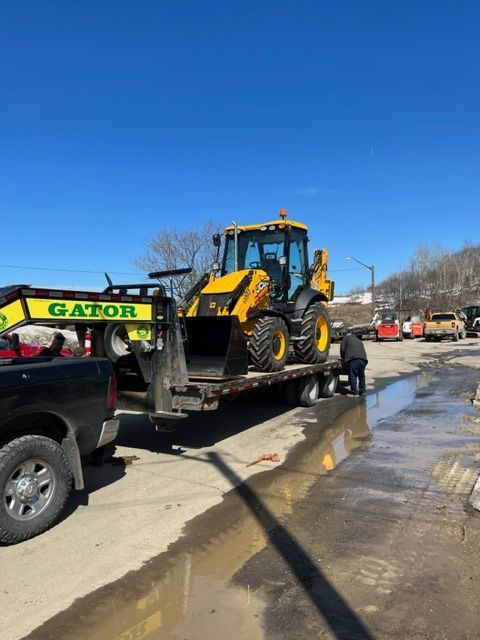 A yellow backhoe on a flatbed trailer being loaded. Black truck, Gator logo, blue sky.