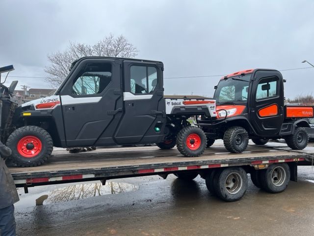 Two Bobcat utility vehicles on a flatbed trailer; one is black and larger, the other orange and smaller.