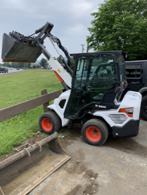 White Bobcat mini loader with orange wheels near a wooden fence and grass. The loader's bucket is raised.