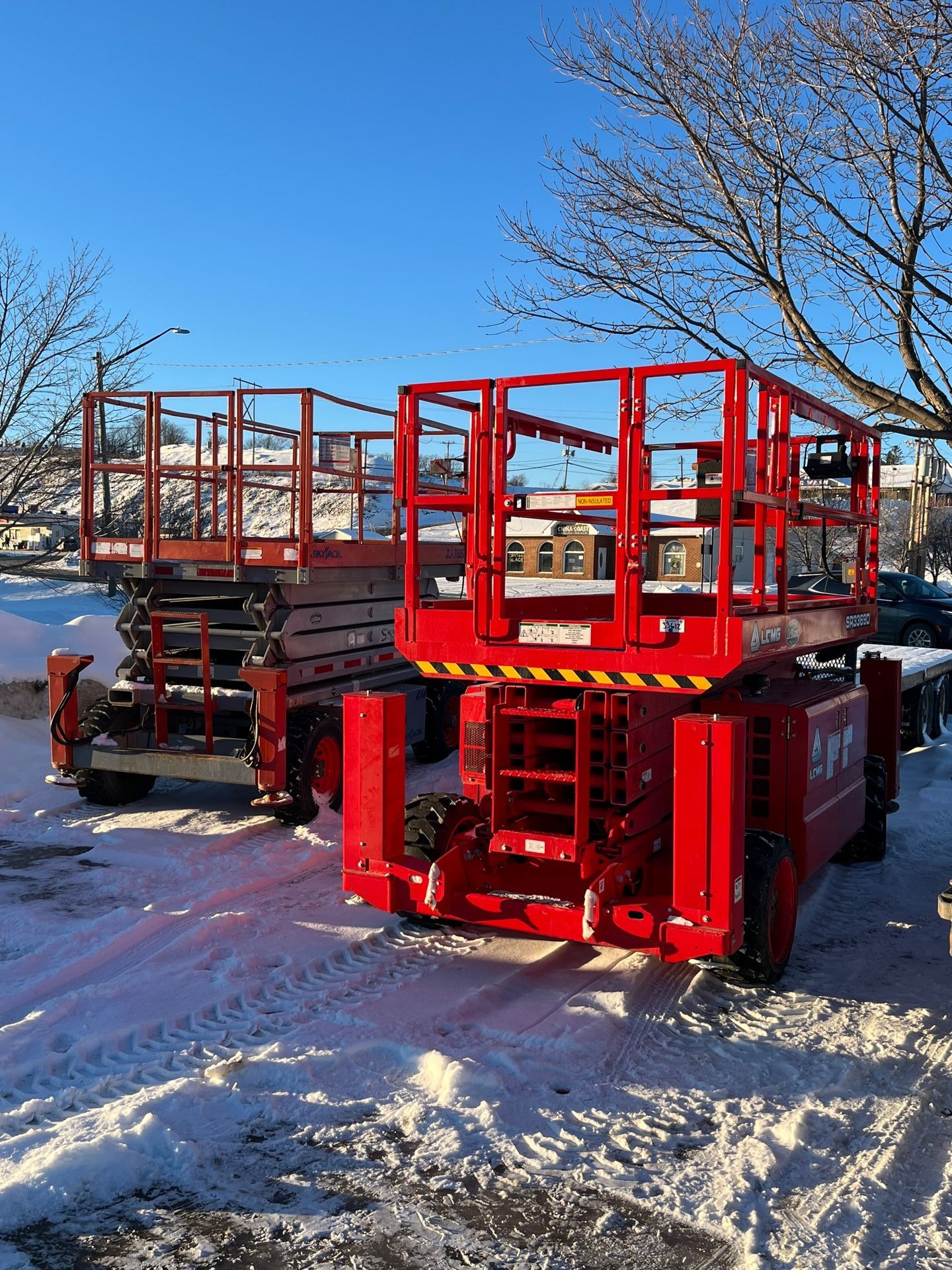 Two red scissor lifts on snow, under a blue sky, near a street.