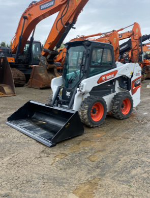 White Bobcat skid-steer loader with a black bucket, orange wheels, and a glass cab on a construction site.