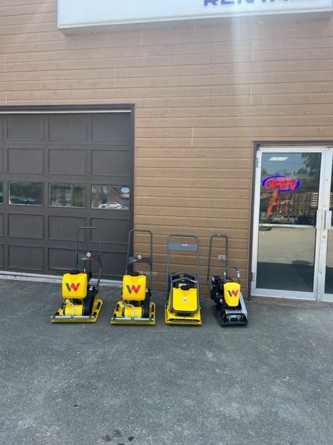 Four yellow Wacker plate compactors lined up in front of a building with a brown garage door.