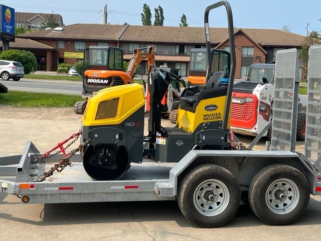 Yellow and black Wacker Neuson compact roller on a trailer, with construction equipment and a building in the background.