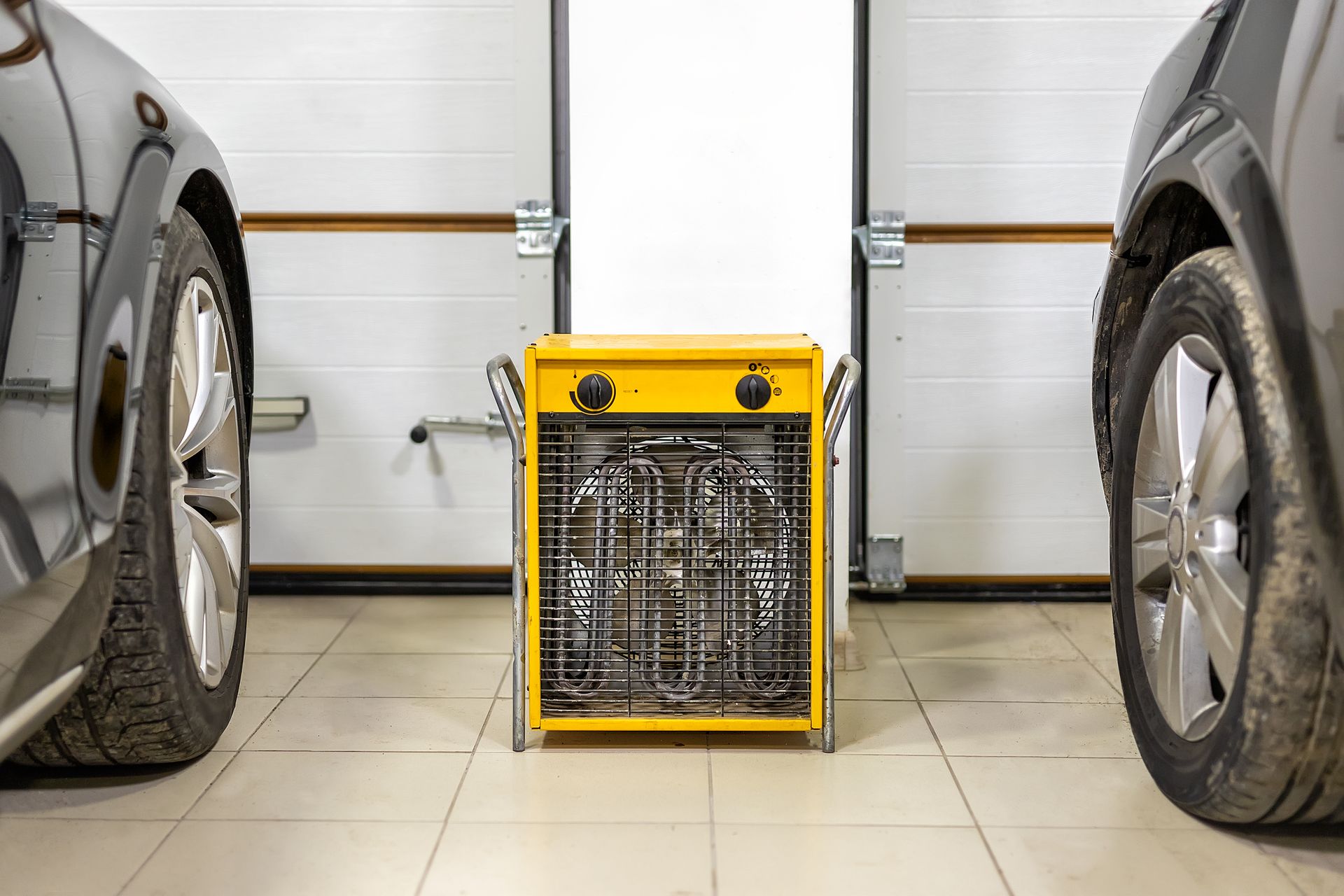 Yellow electric garage heater between two cars, in front of a closed garage door.