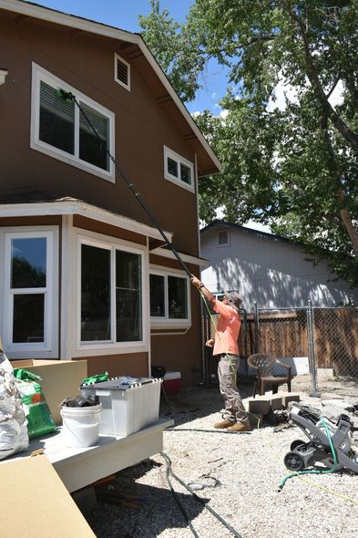 A person in an orange shirt uses a long pole to wash an upper-story window of a brown house.