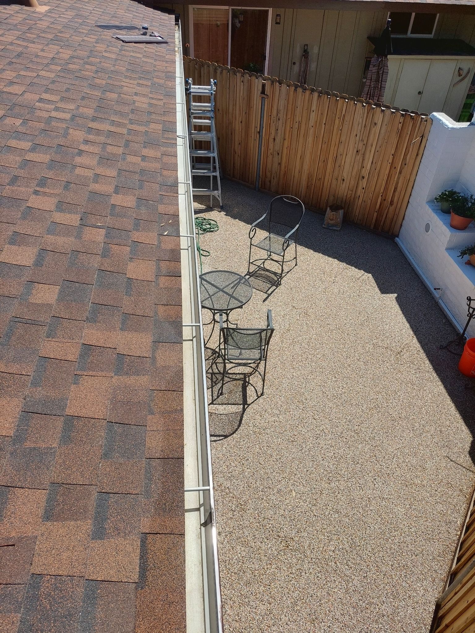 A high-angle view of a backyard with a gravel ground, patio furniture, a wood fence, and the edge of a shingled roof.