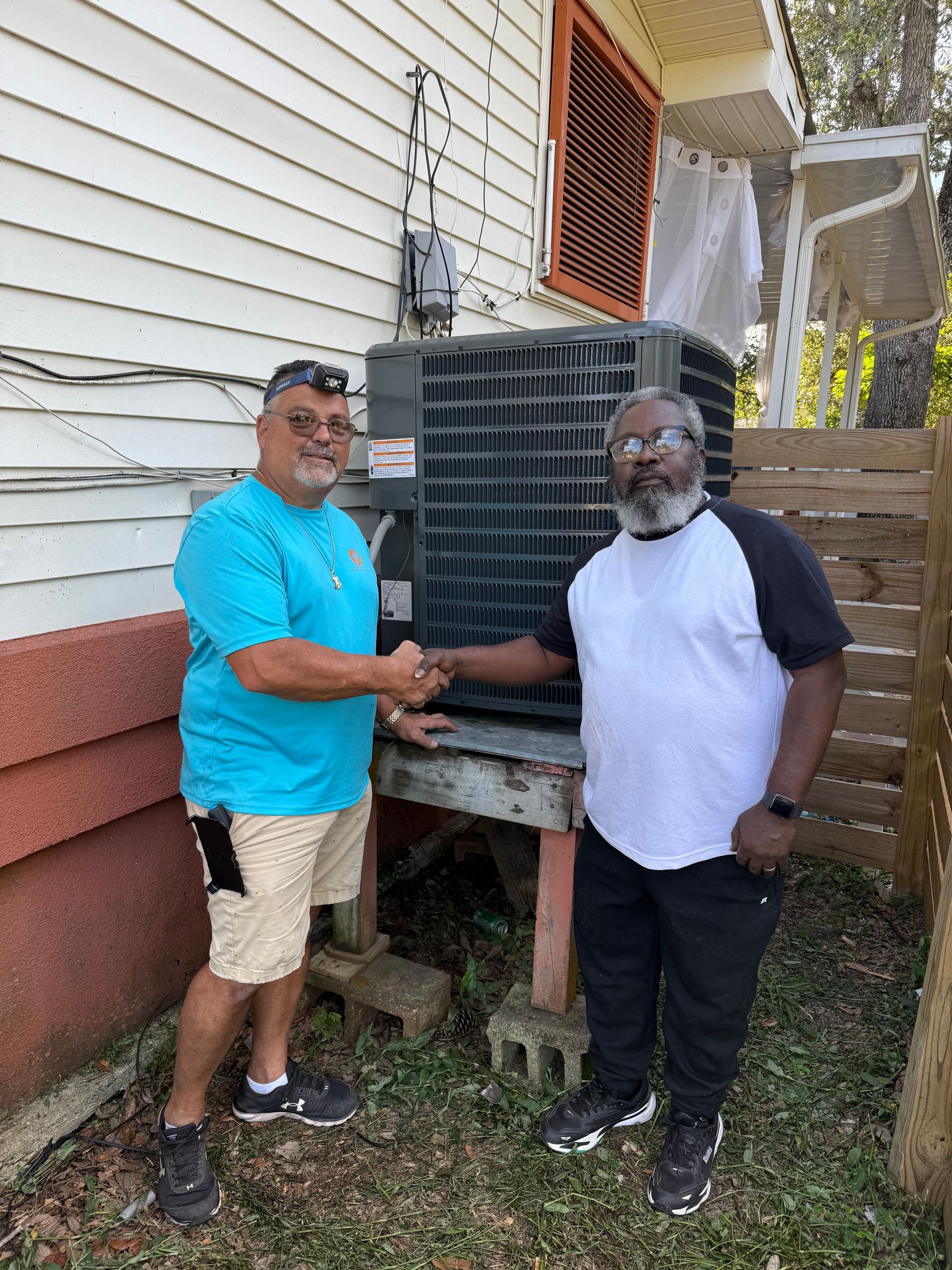 Two men shake hands beside an outdoor air conditioning unit next to a house.
