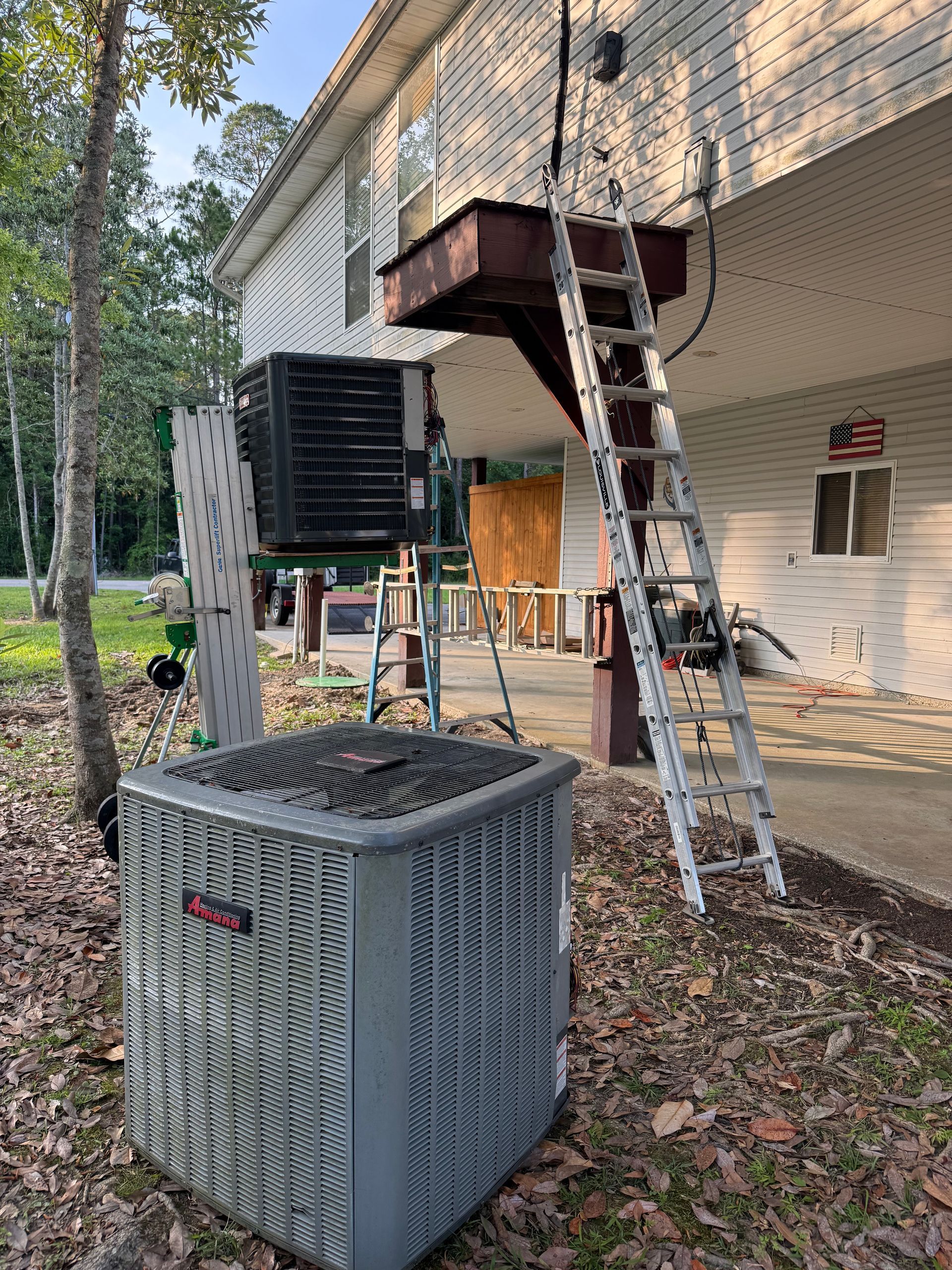 Outdoor scene with an HVAC unit, ladder, and balcony on a beige house in a wooded yard