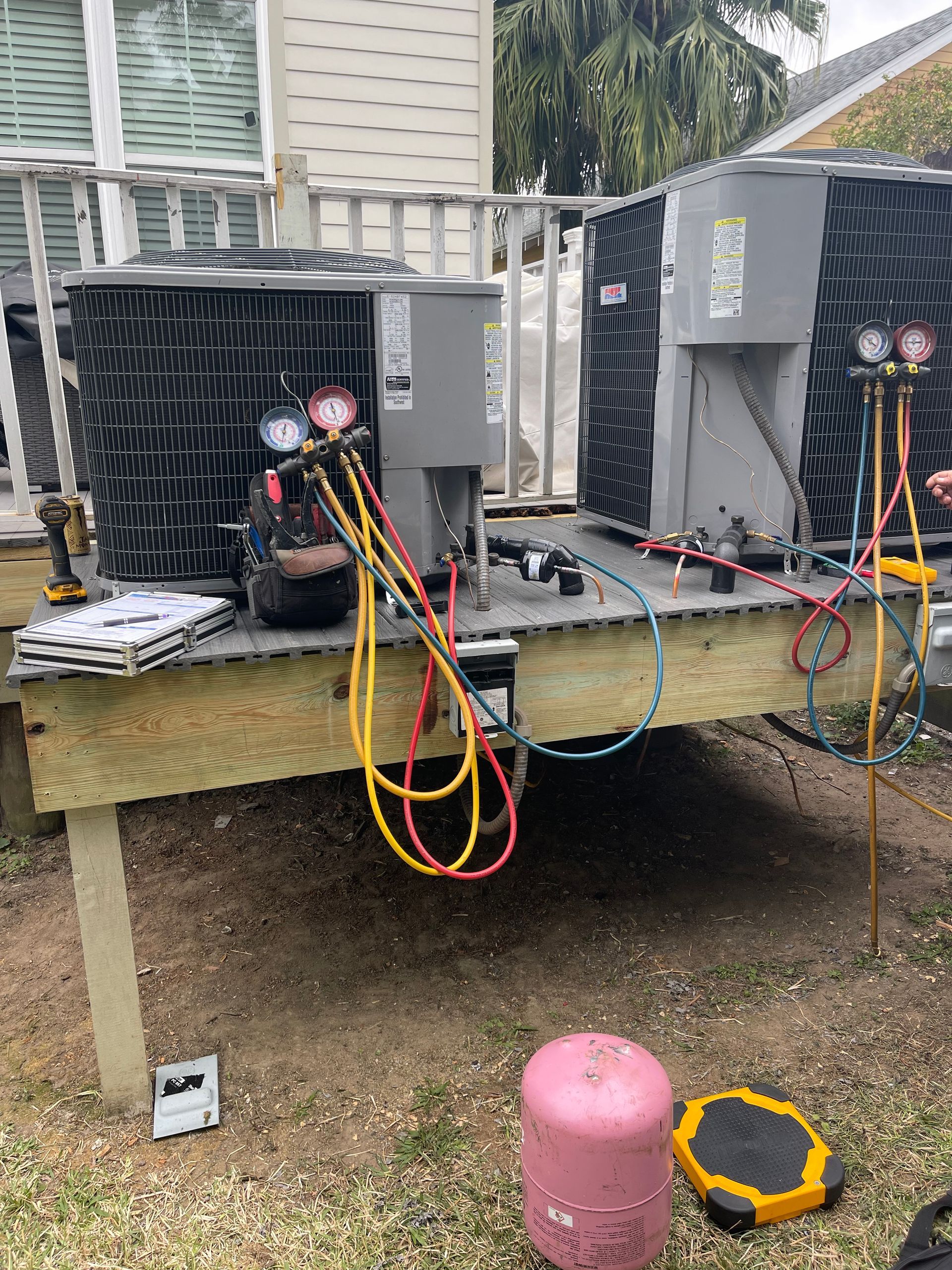 Outdoor HVAC units on a raised platform with colorful service hoses attached, beside a house.