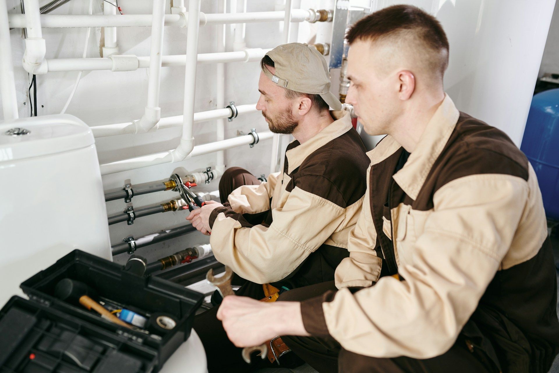Two technicians inspect pipes and tools in a mechanical room with white equipment.