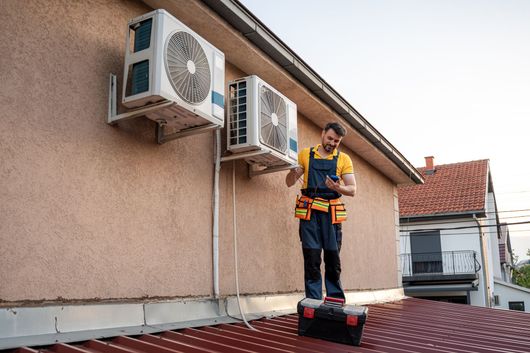 Technician standing on rooftop beside two wall-mounted air conditioning units, holding a tool bag