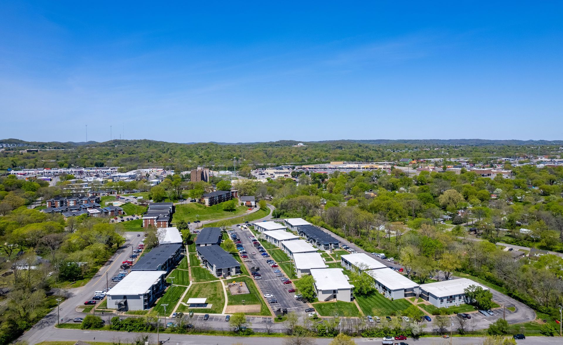 Aerial view of a residential complex with multiple white-roofed buildings surrounded by trees under a clear blue sky.