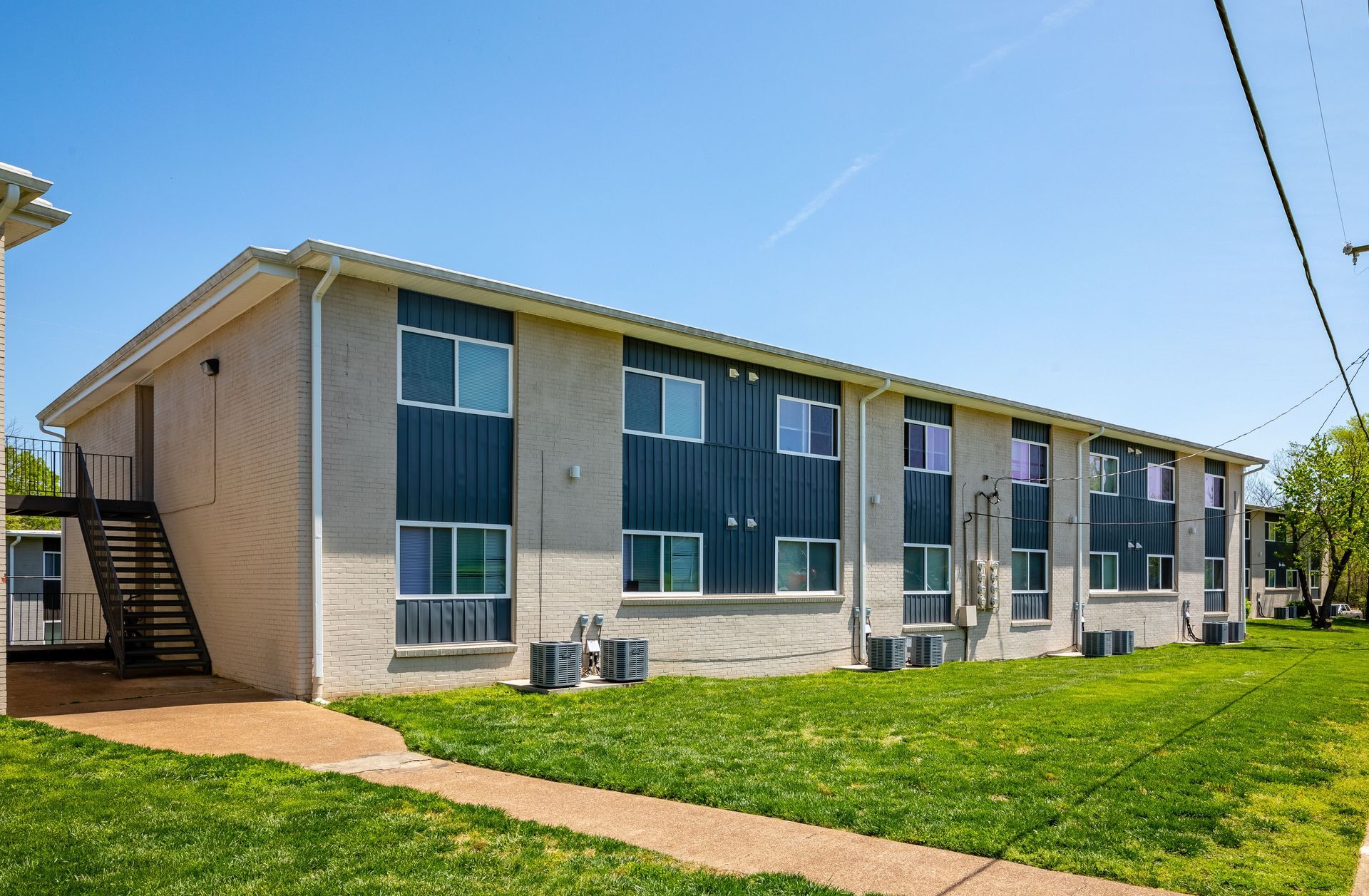 A two-story apartment building with tan stucco, blue accents, and an exterior metal staircase under a clear blue sky.