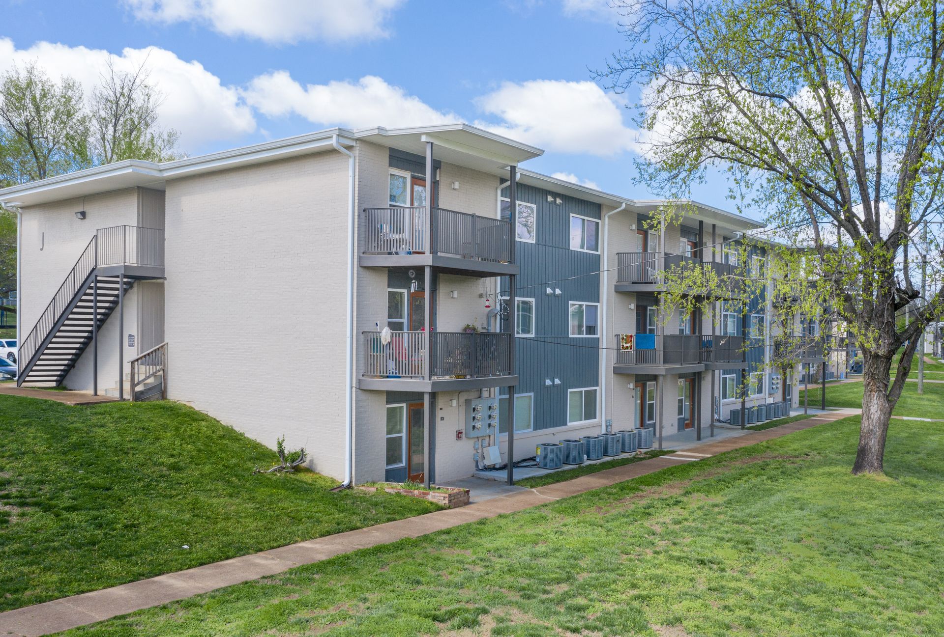 A multi-story apartment building with beige walls and dark gray balconies, set against a lawn under a blue, cloudy sky.