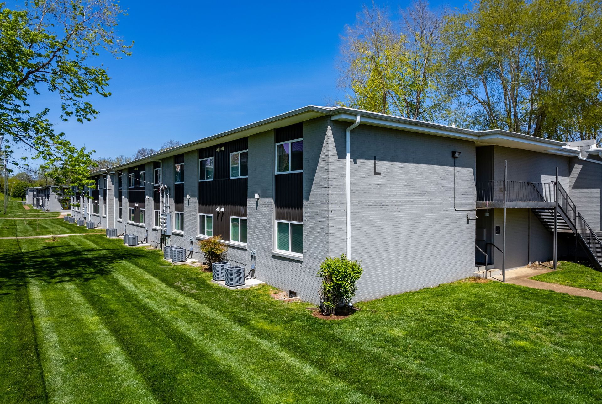 A two-story grey apartment building with a row of air conditioning units, surrounded by a mown green lawn under a blue sky.