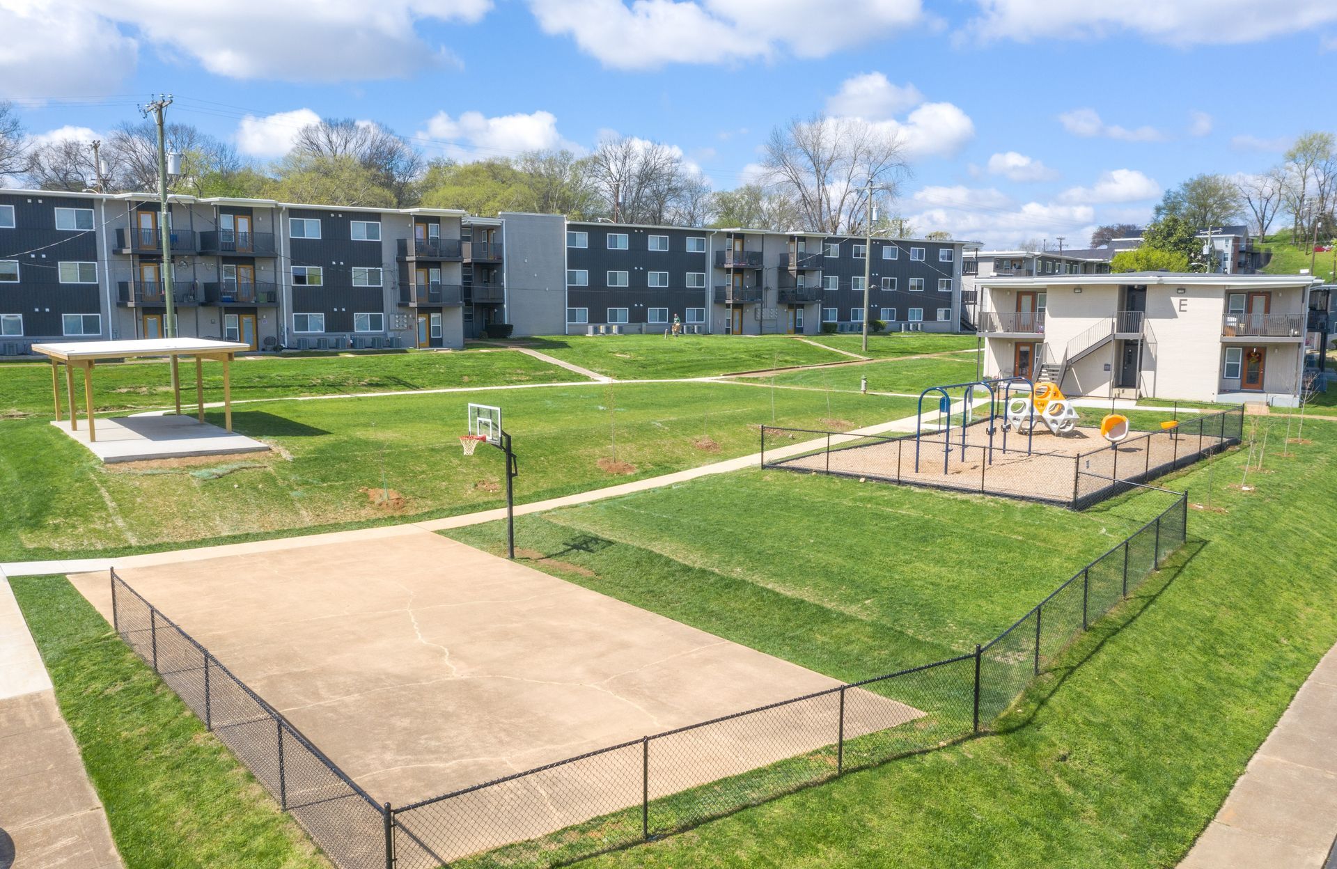 An aerial view of an apartment complex courtyard featuring a fenced basketball court, playground, and shaded seating.