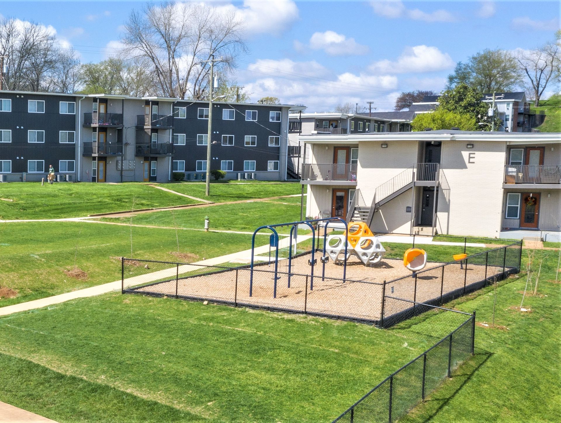 A fenced playground with a play structure sits on a grassy lawn between two multi-story apartment buildings.
