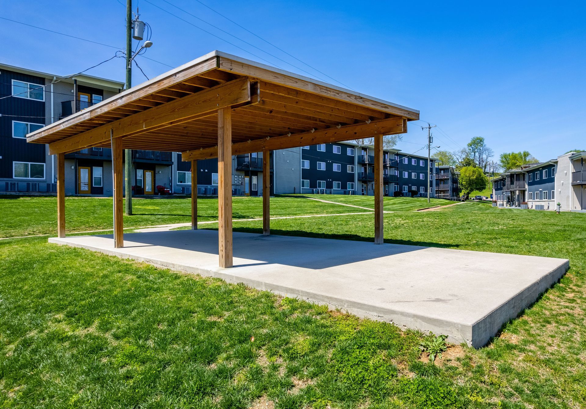 A wooden pavilion with a metal roof stands on a concrete slab in a grassy courtyard between apartment buildings.