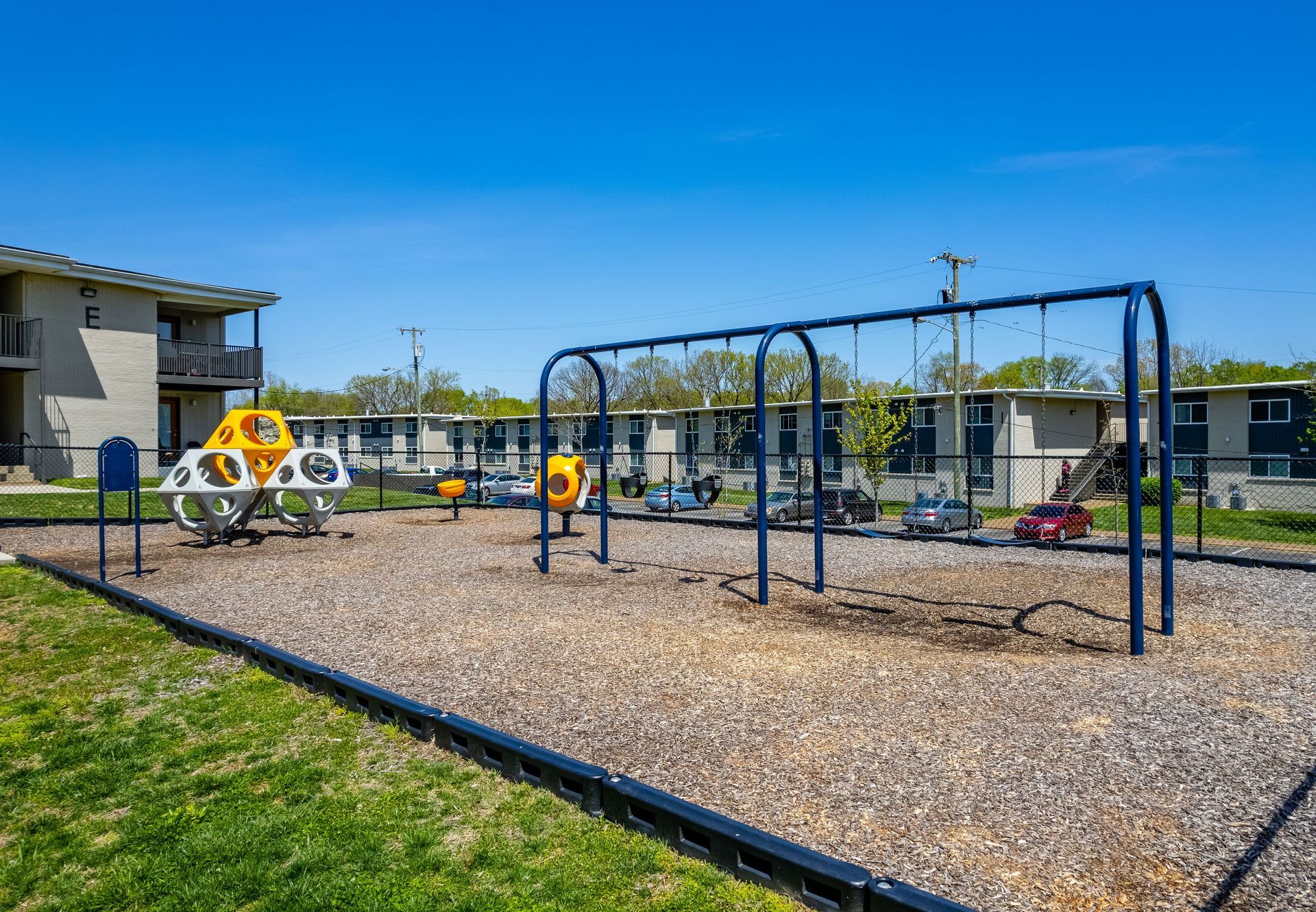 A playground with a blue swing set and a white and yellow climbing structure on wood chips against a clear blue sky.