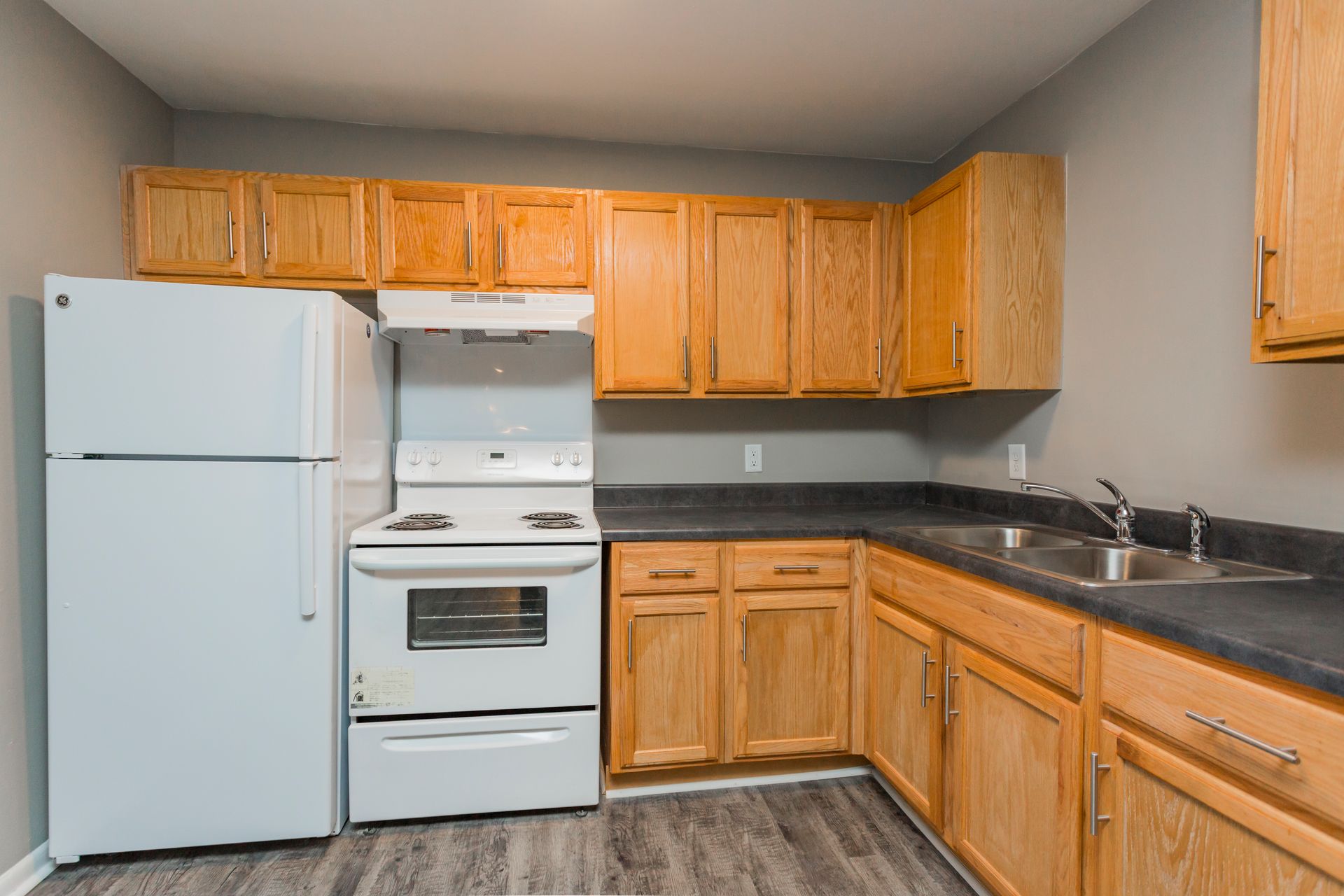 A kitchen with light wood cabinets, gray counters, a white refrigerator, and a white stove, against a light gray wall.