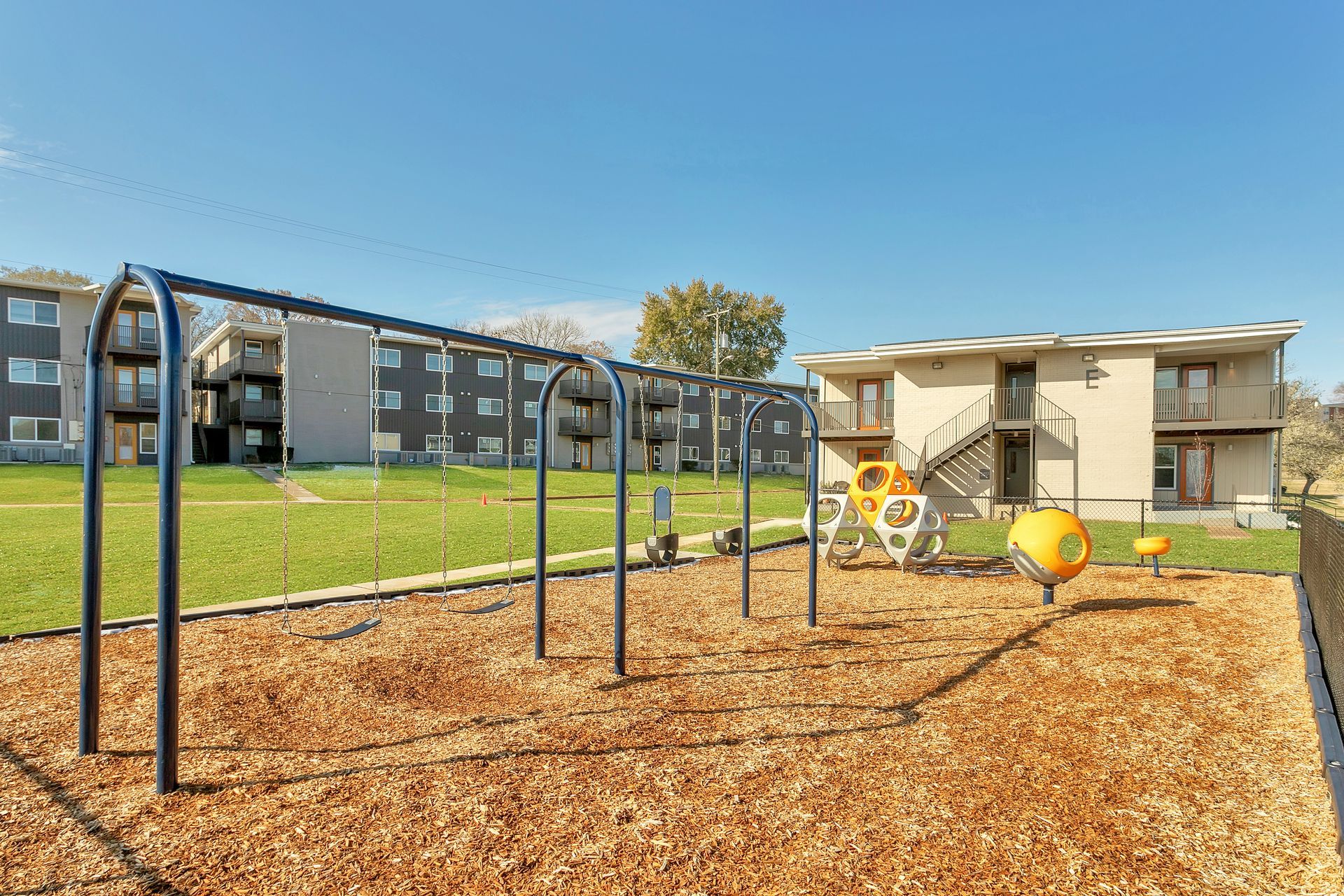 A playground with a swing set and yellow equipment on wood chips in front of apartment buildings under a clear blue sky.
