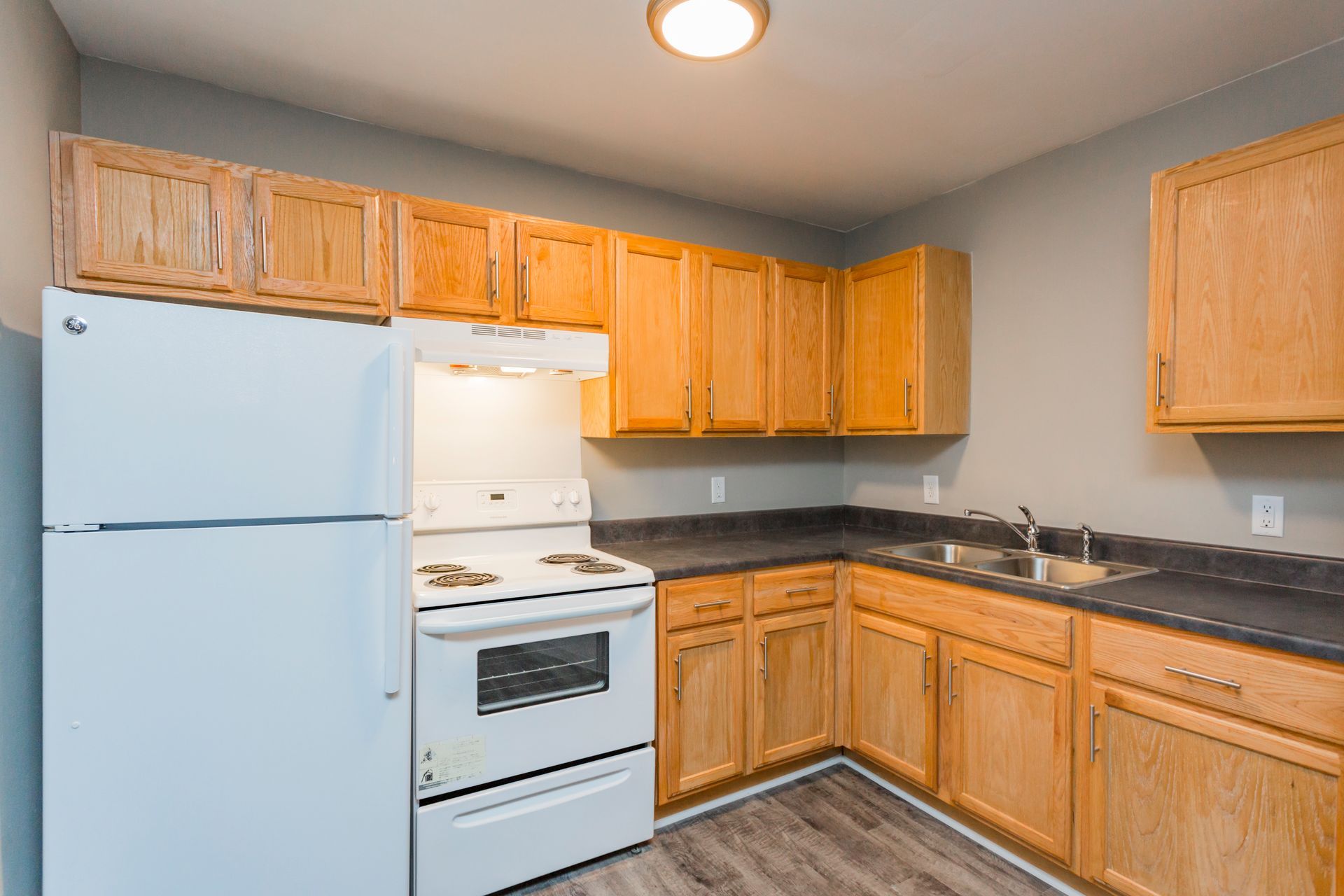 A kitchen with wood cabinets, dark countertops, a white refrigerator, and a white oven, illuminated by a ceiling light.