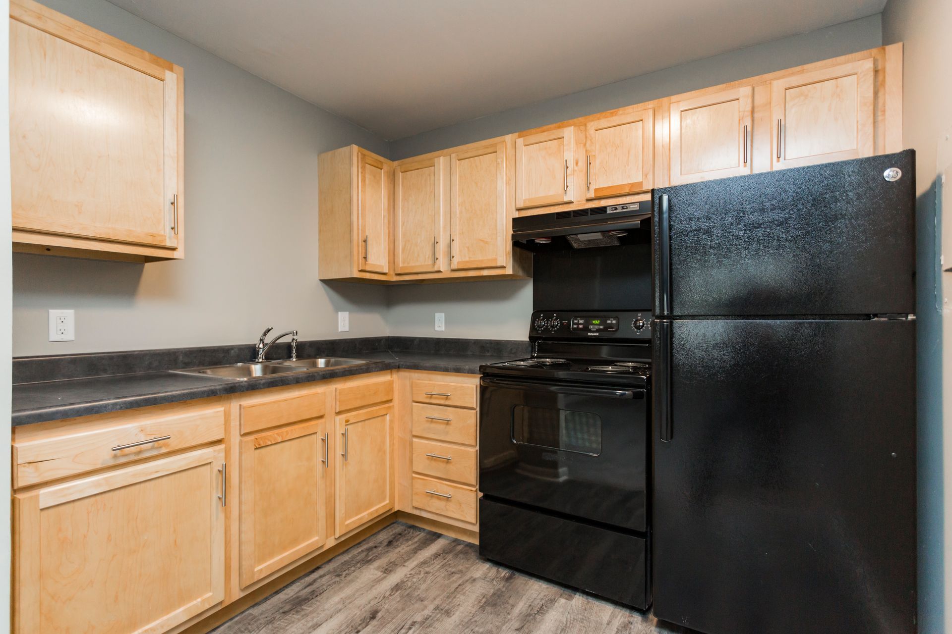 A kitchen with light wood cabinets, dark countertops, a black stove, and a black refrigerator against a gray wall.