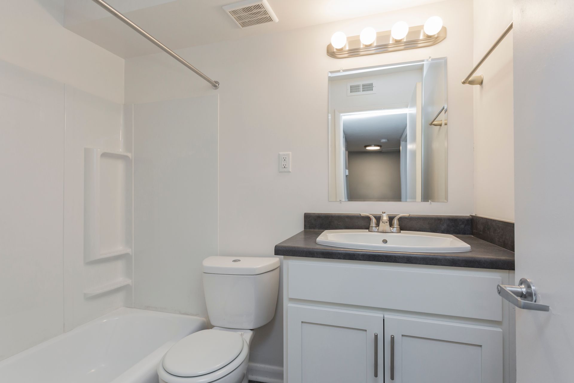 A clean, white bathroom featuring a tub-shower combination, a toilet, and a vanity with a dark countertop and mirror.