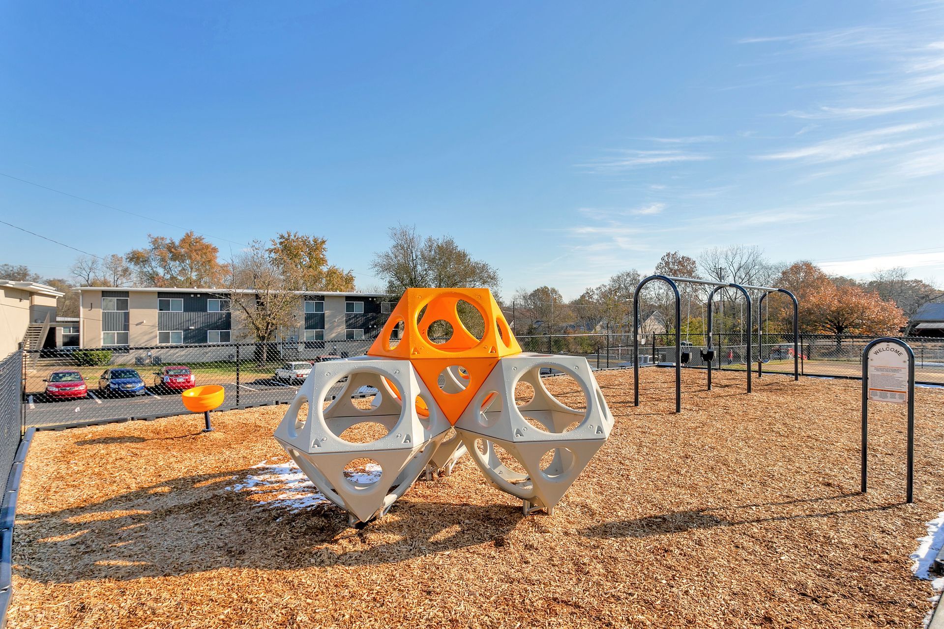 A geometric orange and white climbing structure sits on wood chips in a sunny outdoor playground near apartment buildings.