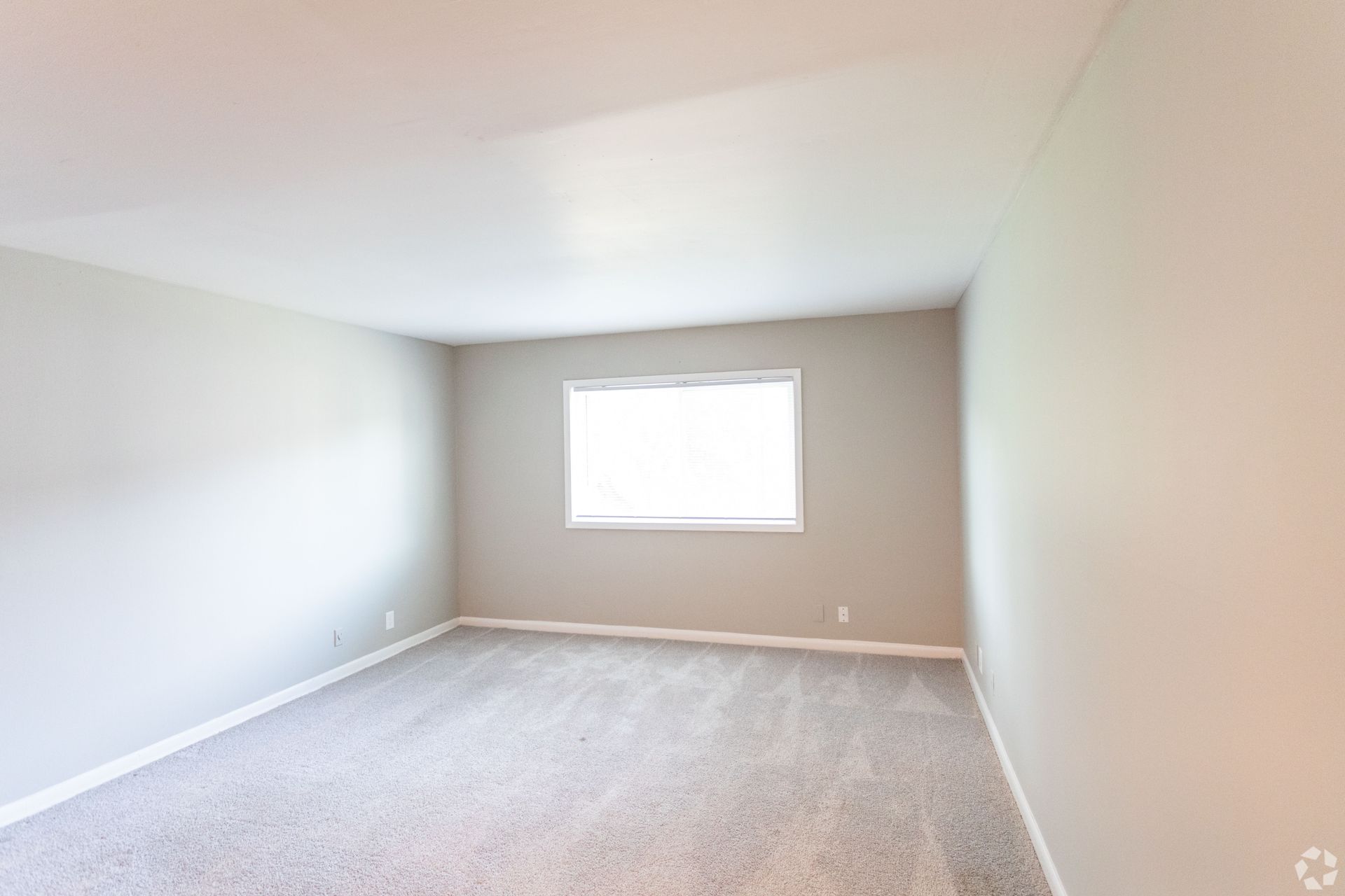An empty bedroom featuring beige walls, light carpet flooring, and a window centered on the far wall.