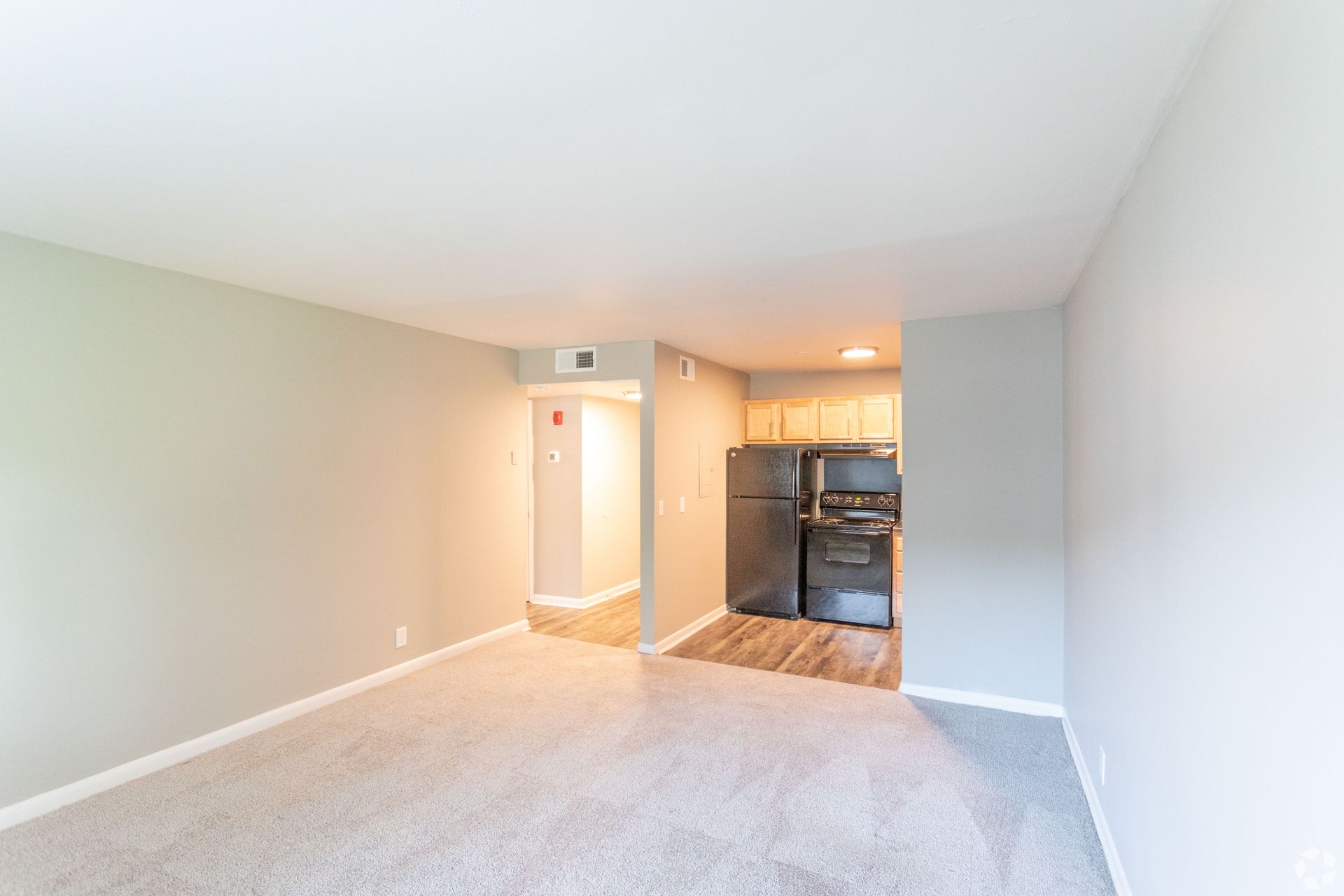 An empty living room with light gray carpet and walls, leading to a small kitchen area with dark appliances and wood floor.