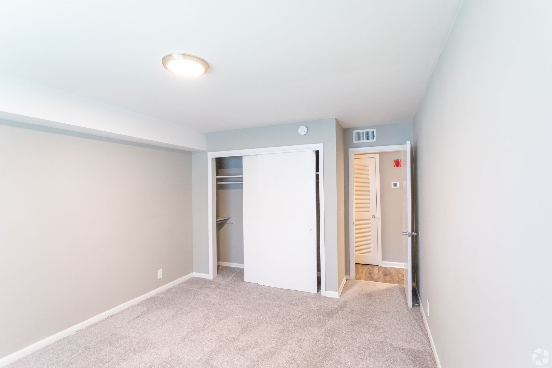 A vacant bedroom with gray walls, beige carpet, a sliding-door closet, and an open doorway leading to a tiled room.